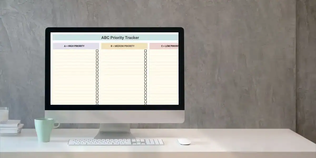 A desktop computer screen displays a colorful Google Sheets task prioritization template titled "ABC Priority Tracker," with three columns labeled A = High Priority, B = Medium Priority, and C = Low Priority, each containing checkbox rows for task management. The workspace includes a minimalist white desk setup with a keyboard, mouse, books, and a mint green coffee mug.