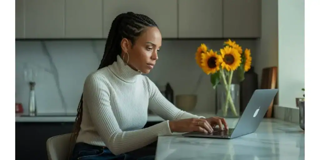 Woman working on a laptop at a kitchen counter with a vase of sunflowers in the background, appearing focused and productive—ideal for promoting a Google Sheets Email Marketing Template designed to streamline campaign planning from home.