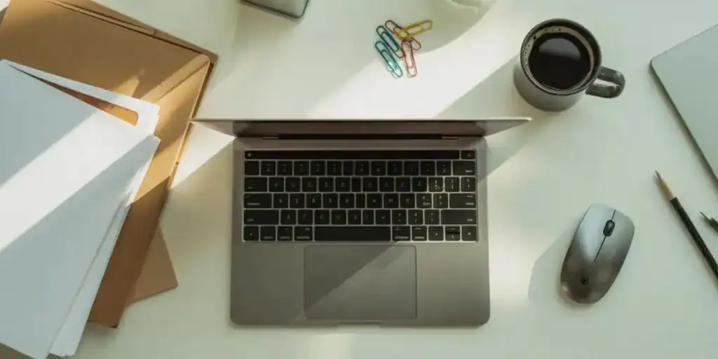 Top-down view of a minimalist desk with an open laptop, coffee mug, wireless mouse, pencil, folders, and colorful paperclips—ideal for organizing workflows or referencing a Google Sheets SOP tracker during daily planning.

