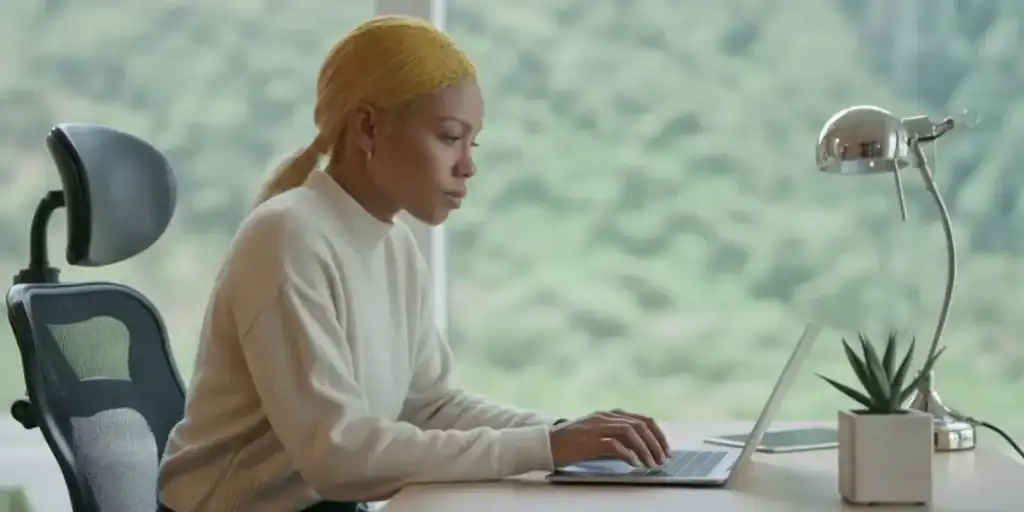 A woman in a light sweater working intently on a laptop at a minimalist desk setup, with a desk lamp and small plant nearby, suggesting she is managing tasks or processes using a Google Sheets SOP tracker in a serene, natural-lit workspace.

